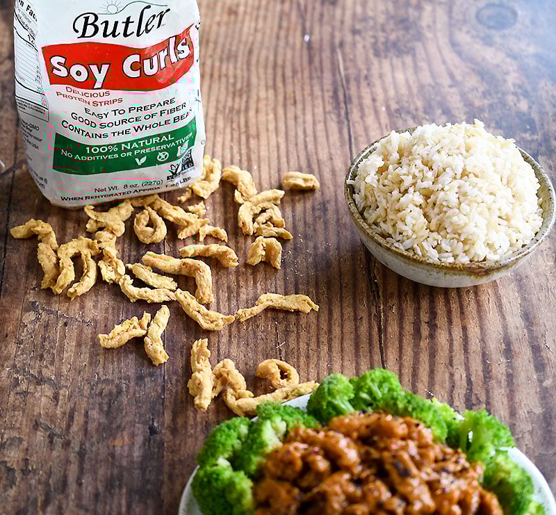 Butler Soy Curls scattered on wooden table with bowl of brown rice and plate of Vegan General Tso