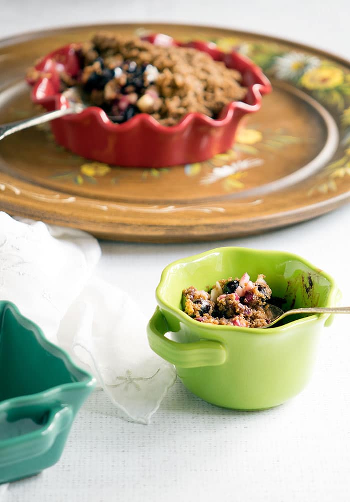 Vegan crumble in a green ramekin with handles on a white tables with a wooden plate with painted flowers on it.