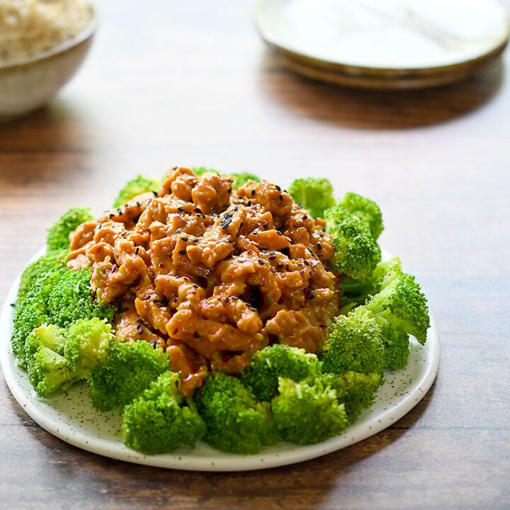 General Tso Soy Curls surrounded by a circle of steamed broccoli on a white ceramic plate on a dark wooden table