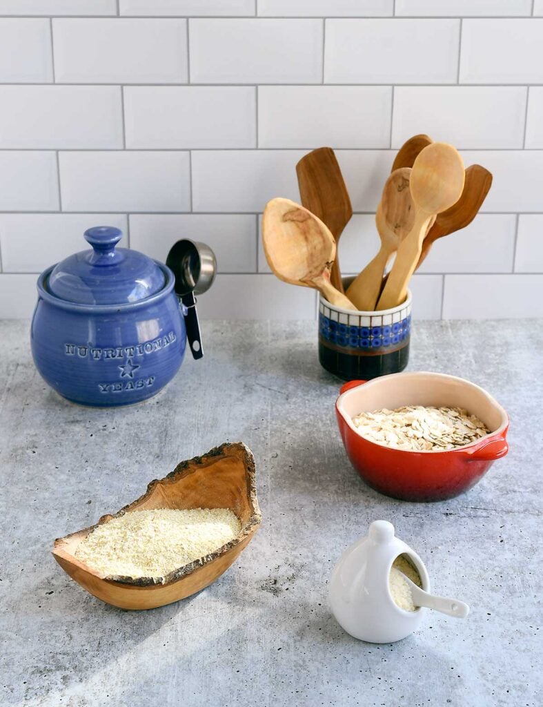 Homemade vegan parmesan in a wooden bowl, with a red ceramic bowl of oats and hand carved wooden spoons.