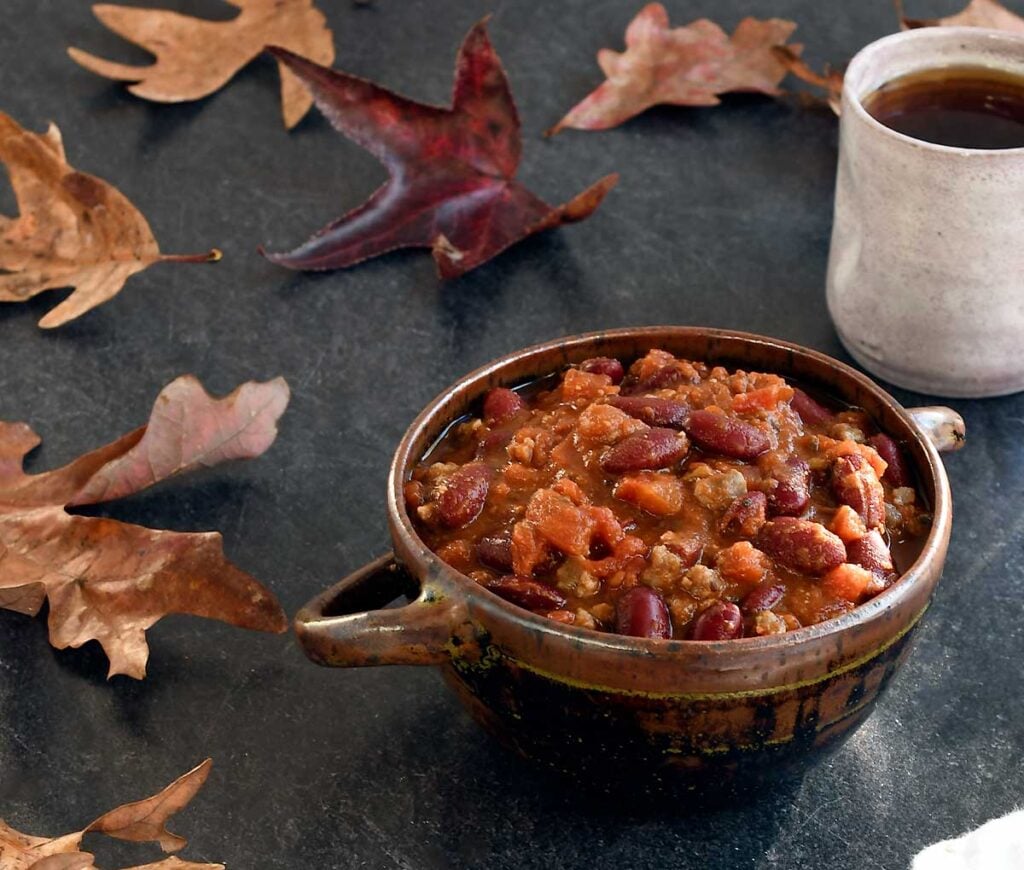 Vegan chili in a ceramic bowl with 2 handles, hot tea, on a slate table with autumn leaves.